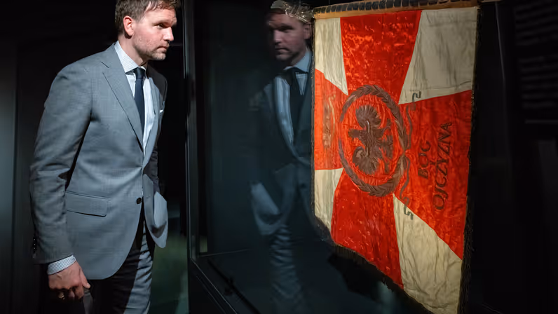 Derk Boswijk, staatssecretaris van Defensie, draagt een grijs pak en staat naast het vaandel in de expositie en kijkt naar de bannier.