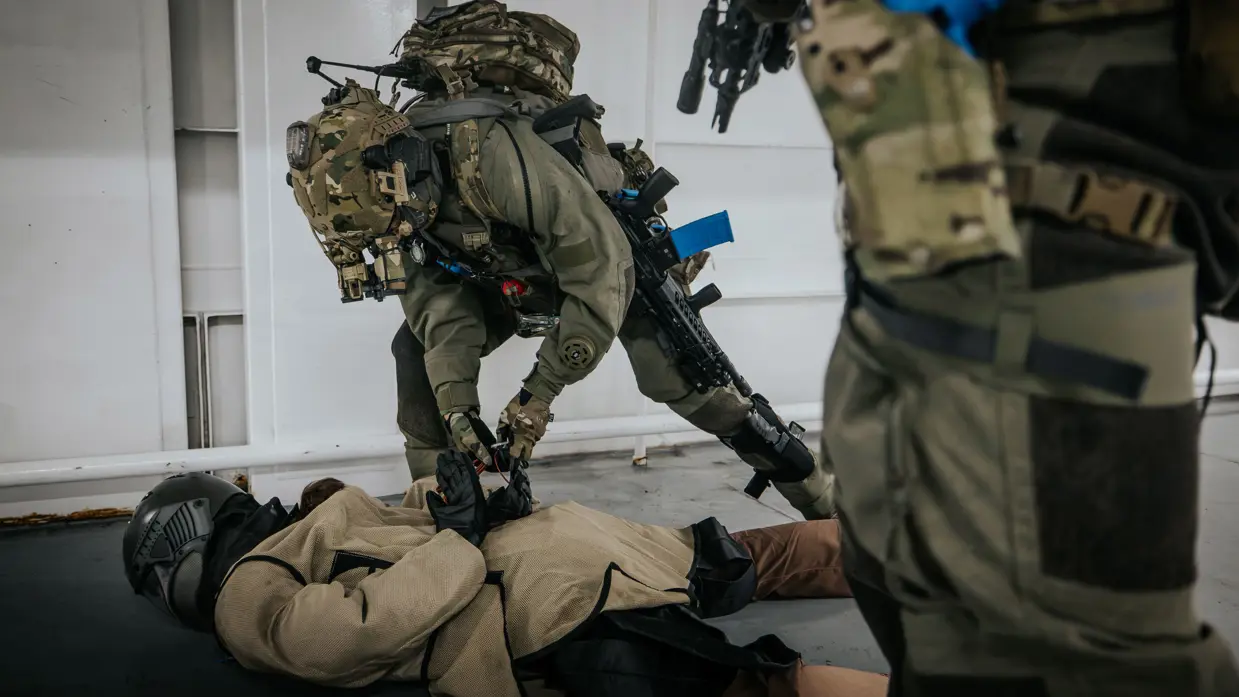 Een NLMARSOF-operator boeit een op de grond liggende oefenvijand. Een collega-operator kijkt toe. De oefenvijand draagt een helm en beschermende kleding. 