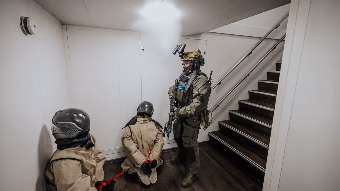 Een operator van NLMARSOF bewaakt twee gevangenen die geknield op de grond zitten, neus richting de muur.