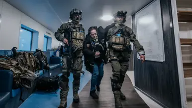 Links controleert een NLMARSOF-militair een vrouw die bleek ziet en duidelijk in nood verkeert. Rechts ondersteunen 2 gewapende NLMARSOF-operators een gewonde dame. Ze lopen over een scheepsdek.