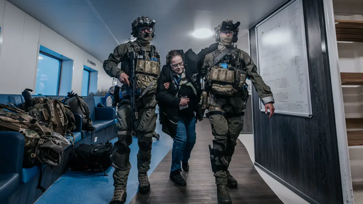 Links controleert een NLMARSOF-militair een vrouw die bleek ziet en duidelijk in nood verkeert. Rechts ondersteunen 2 gewapende NLMARSOF-operators een gewonde dame. Ze lopen over een scheepsdek.