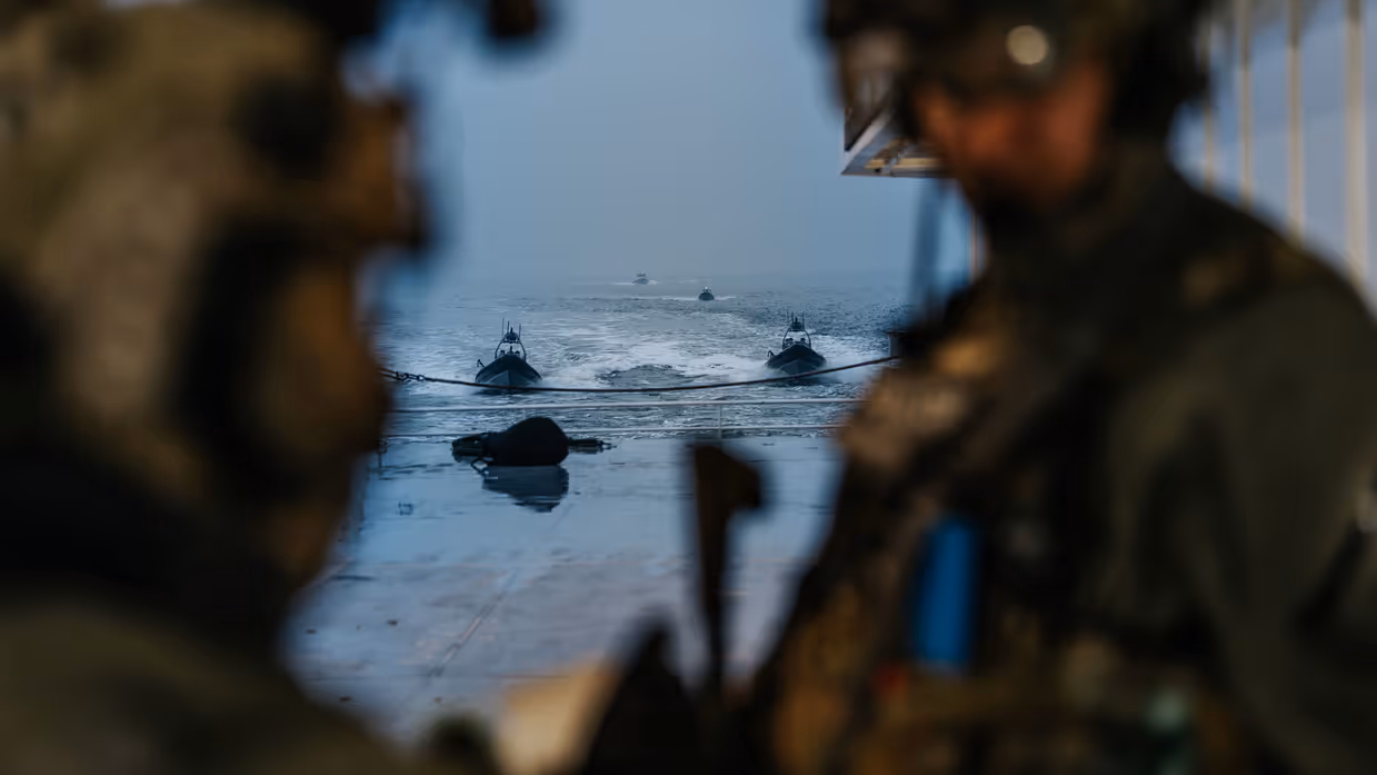 Links staan 2 NLMARSOF-operators  op de voorgrond, op de achtergrond zijn snelle onderscheppingsvaartuigen te zien die achter de veerboot varen. Rechts kijkt een gemaskerde NLMARSOF-operator om een hoekje, wapen gereed.