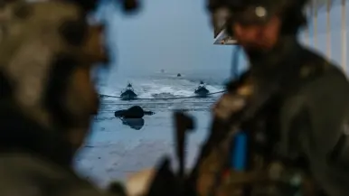 Links staan 2 NLMARSOF-operators  op de voorgrond, op de achtergrond zijn snelle onderscheppingsvaartuigen te zien die achter de veerboot varen. Rechts kijkt een gemaskerde NLMARSOF-operator om een hoekje, wapen gereed.