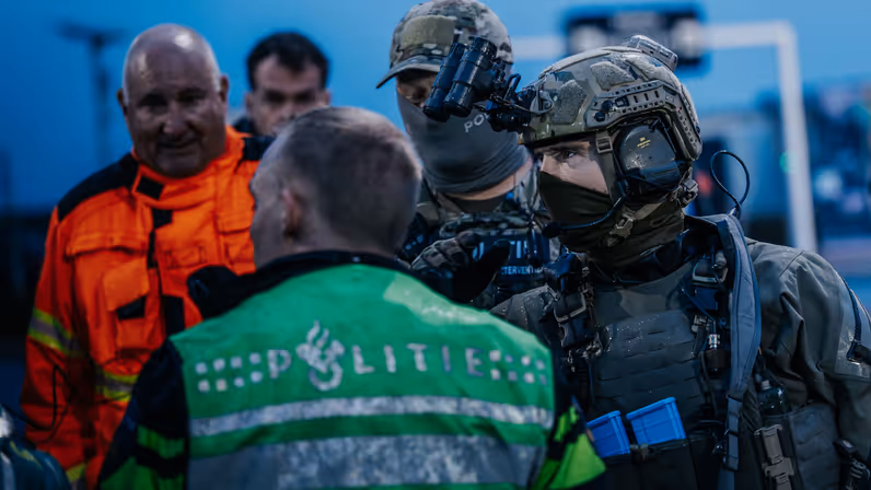 Een gemaskerde NLMARSOF-militair overlegt met een politieagent. Op de achtergrond kijkt een andere hulpverlener in oranje pak toe.