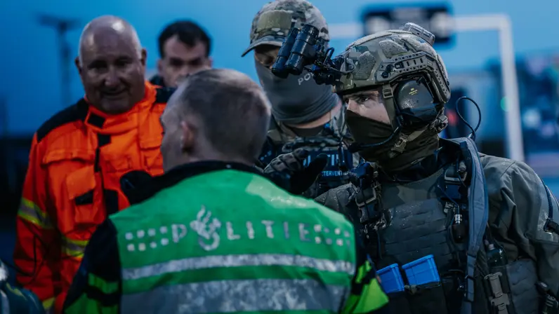 Een gemaskerde NLMARSOF-militair overlegt met een politieagent. Op de achtergrond kijkt een andere hulpverlener in oranje pak toe.