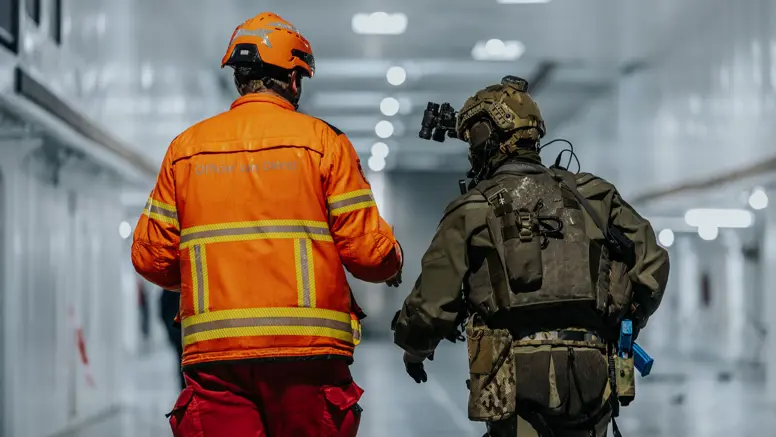 Alt: Twee figuren op de rug gezien, lopend door een verlicht scheepsruim. De linker heeft een fel oranje pak aan en helm op, de rechter is militair en draagt legergroen. 