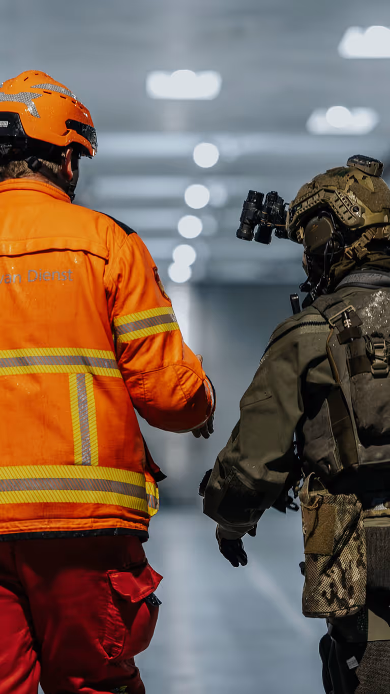 Alt: Twee figuren op de rug gezien, lopend door een verlicht scheepsruim. De linker heeft een fel oranje pak aan en helm op, de rechter is militair en draagt legergroen. 