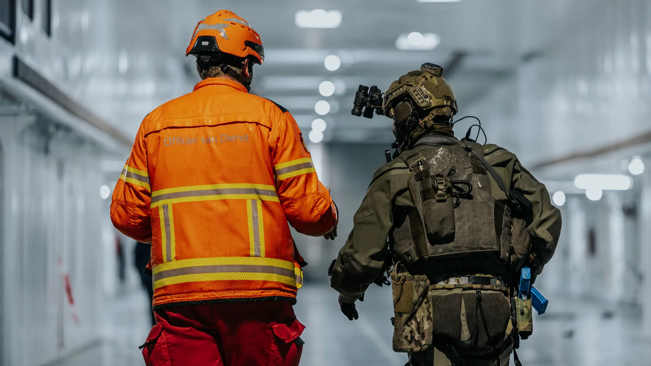 Alt: Twee figuren op de rug gezien, lopend door een verlicht scheepsruim. De linker heeft een fel oranje pak aan en helm op, de rechter is militair en draagt legergroen. 