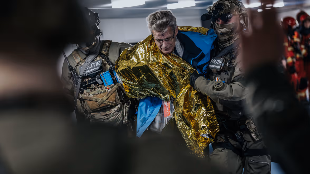 Tijdens een evacuatie heeft een gewonde man een gouden deken omgeslagen, terwijl twee operators hem ondersteunen. 