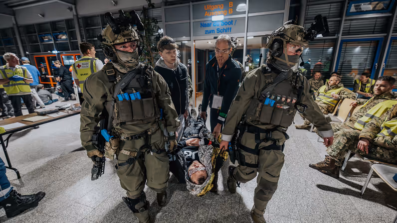 Mariniers tillen met enkele mannen een gewond persoon. Zij komen binnen in een hal vol mensen en hulpverleners. 