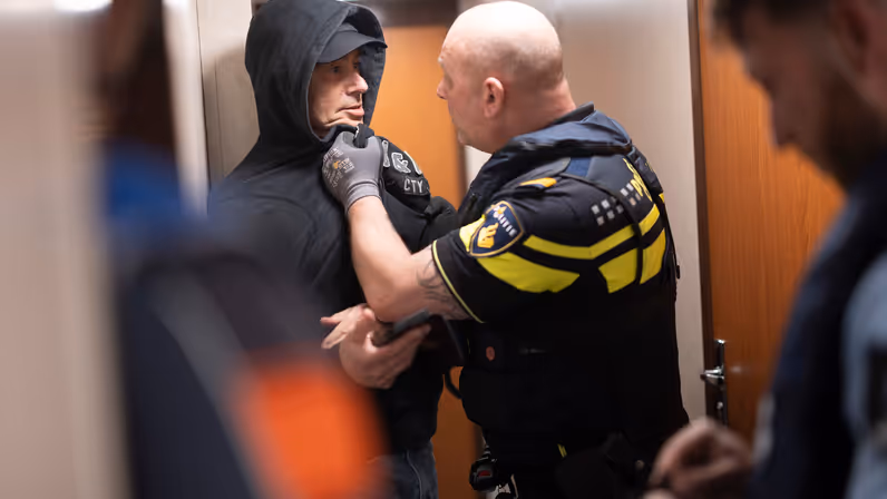 Een kale politieman grijpt een man met pet en capuchon vast.