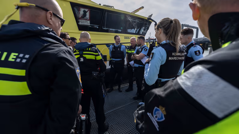 Marechaussees en politiemensen praten met elkaar op het dek van een schip.