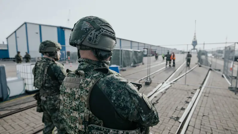 2 militairen, vanaf de rug bezien, bewaken het Tijdelijk Militair Object. Ze kijken naar de hekken voor hen. 