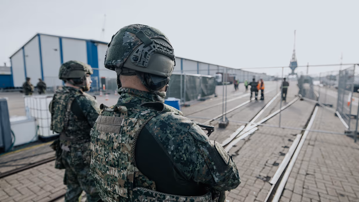 2 militairen, vanaf de rug bezien, bewaken het Tijdelijk Militair Object. Ze kijken naar de hekken voor hen. 