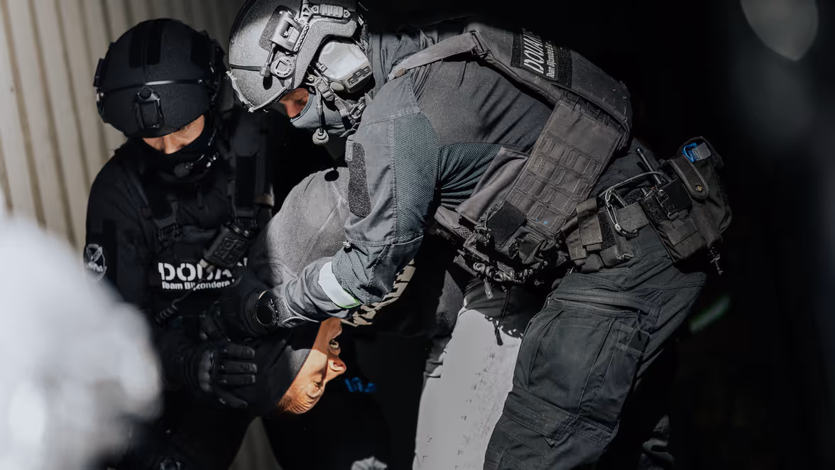 Twee douaniers in donkere pakken dragen een man met zich mee, achter hen is een container te zien.