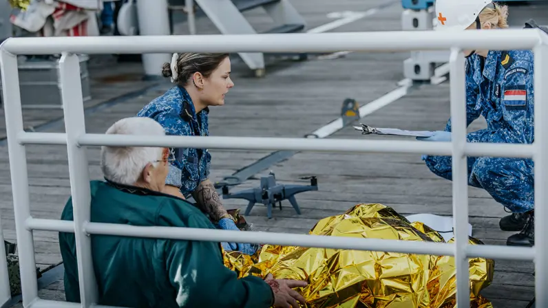 2 militairen in boordtenue behandelen een gewonde man. Hij is in een gouden folie omwikkeld. Op de achtergrond is een drone te zien. 