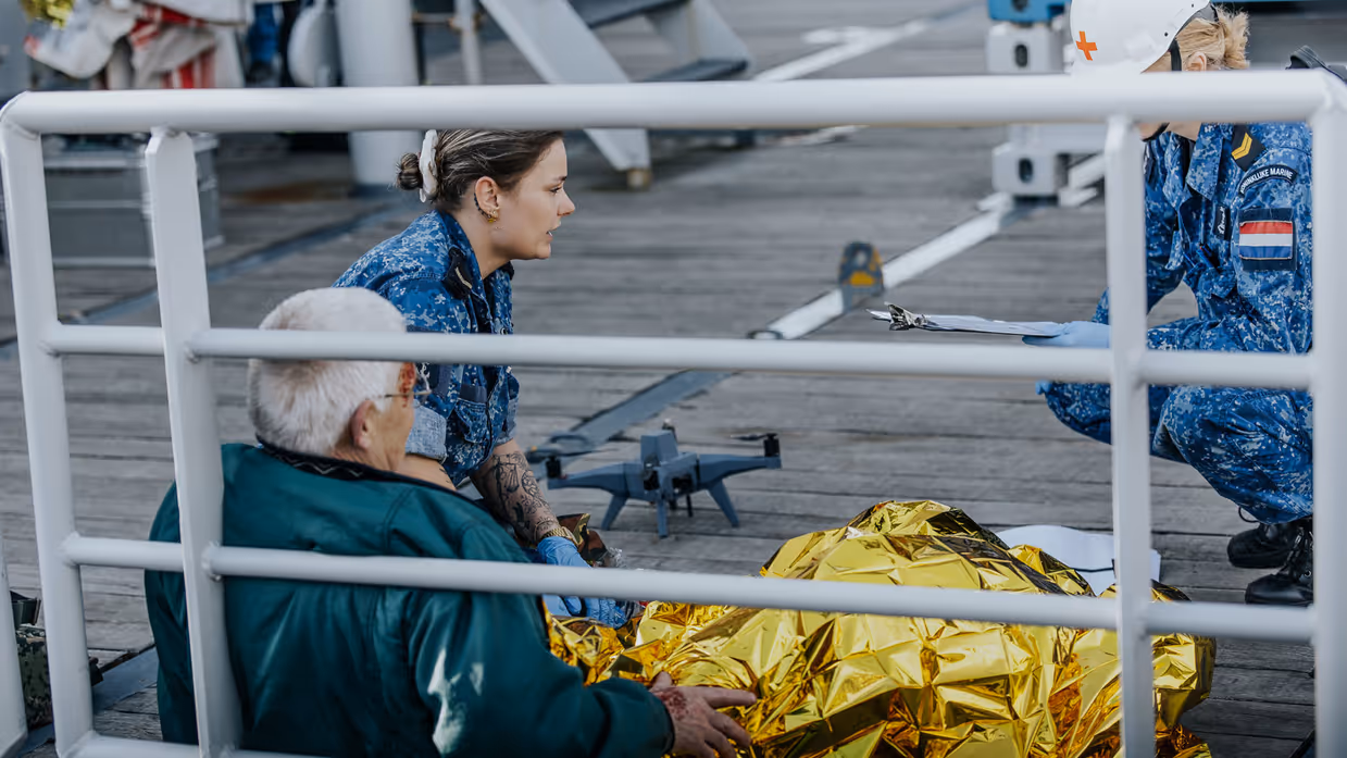 2 militairen in boordtenue behandelen een gewonde man. Hij is in een gouden folie omwikkeld. Op de achtergrond is een drone te zien. 