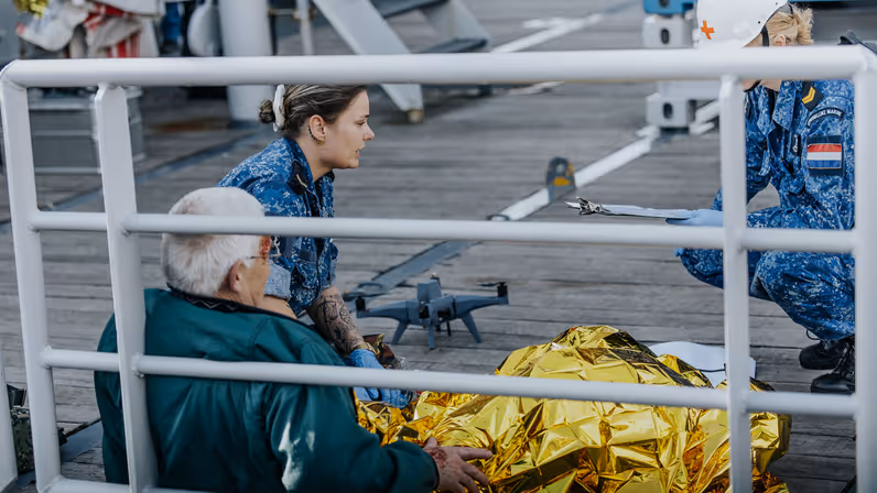 2 militairen in boordtenue behandelen een gewonde man. Hij is in een gouden folie omwikkeld. Op de achtergrond is een drone te zien. 