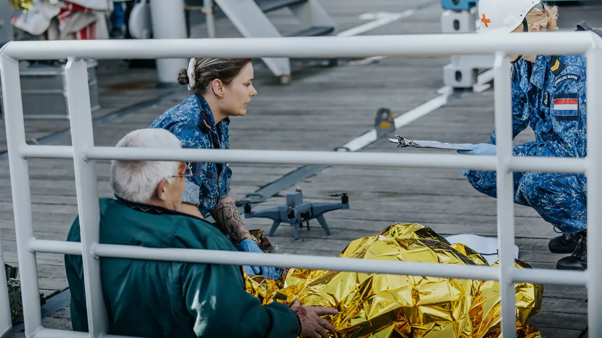 2 militairen in boordtenue behandelen een gewonde man. Hij is in een gouden folie omwikkeld. Op de achtergrond is een drone te zien. 