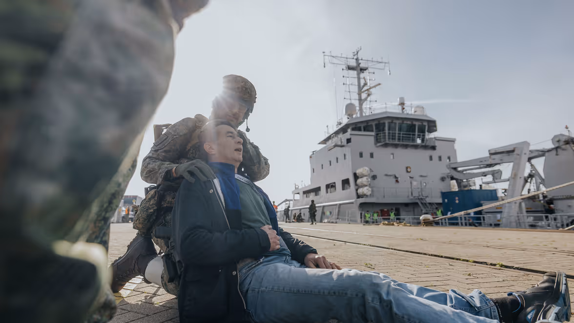 Een militair in groen vlekkenpak ondersteunt een gewonde burger. Op de achtergrond is Zr.Ms. Luymes te zien, aangemeerd aan de kade.  