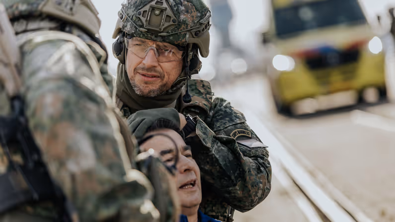  Een militair houdt het hoofd vast van een gewonde man. Op de achtergrond is een ambulance te zien.&nbsp;
