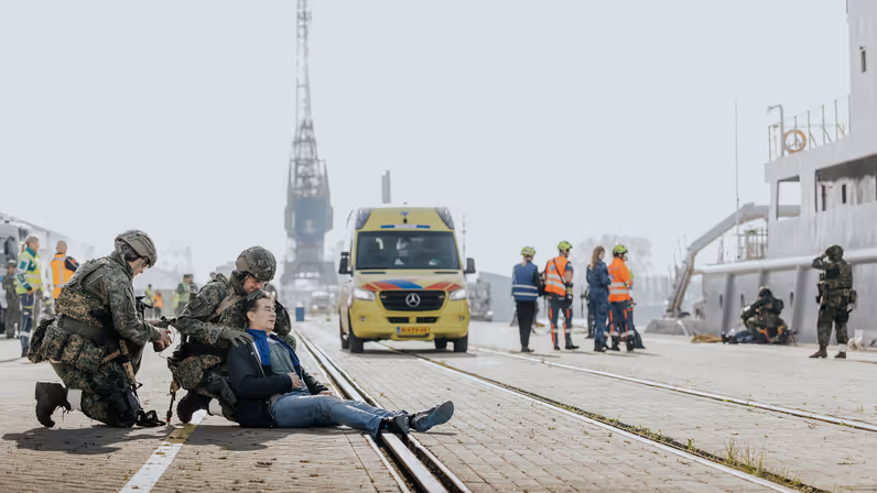 Een militair behandelt een gewonde man, die op de kade ligt. Op de achtergrond is Zr.Ms. Luymes te zien. 