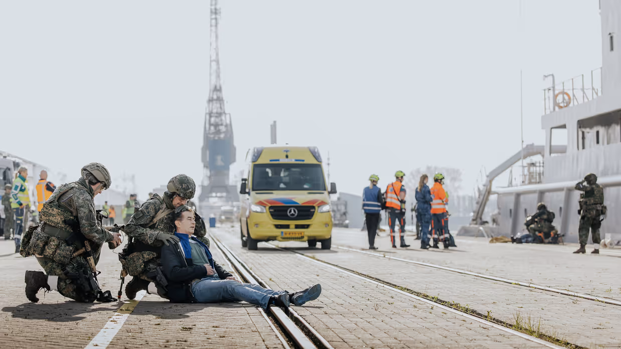 Een militair behandelt een gewonde man, die op de kade ligt. Op de achtergrond is Zr.Ms. Luymes te zien. 