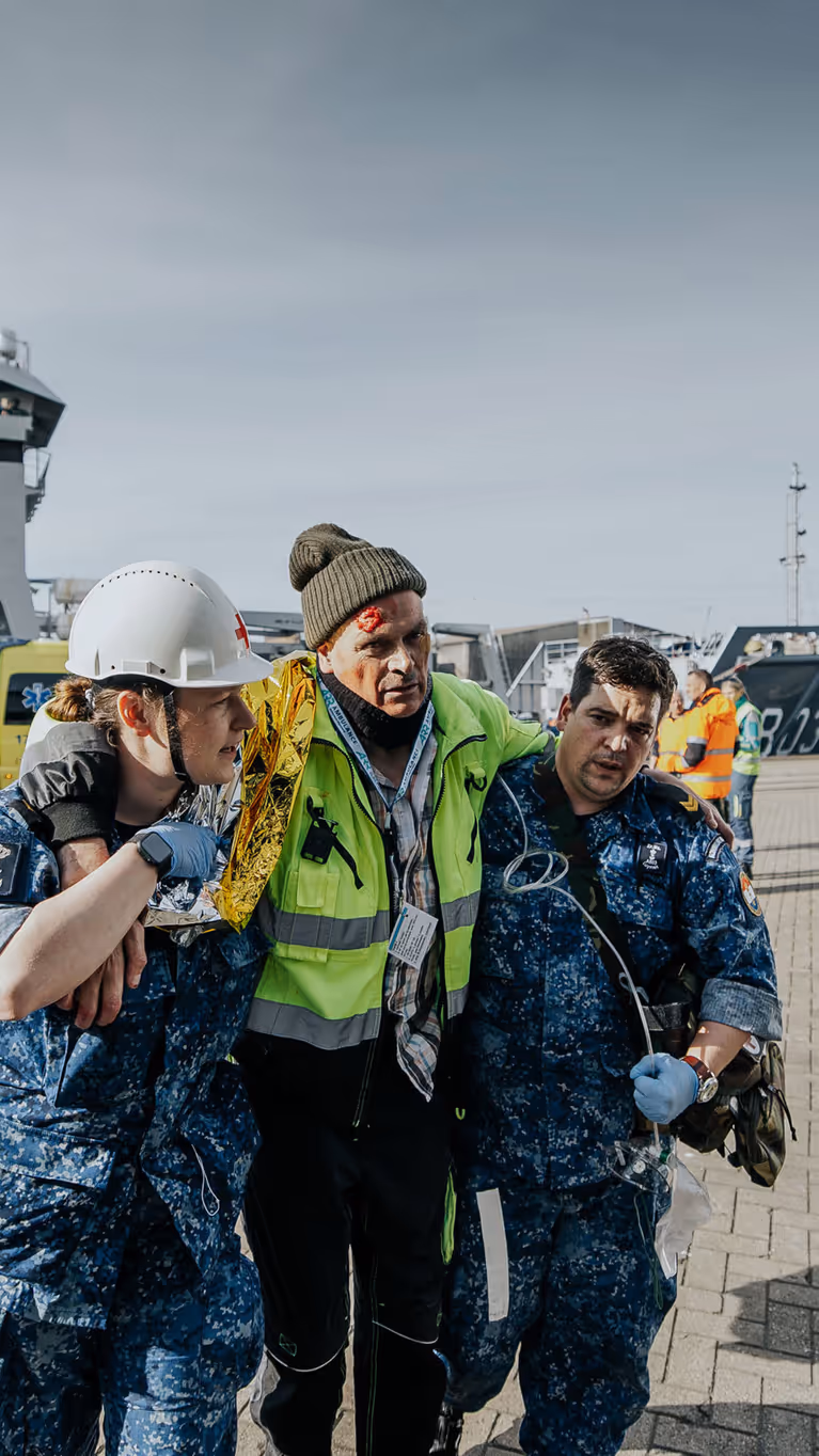 Een gewonde man wordt bij het lopen ondersteund door 2 militairen in boordtenue. Op de achtergrond is Zr.Ms. Luymes te zien.  