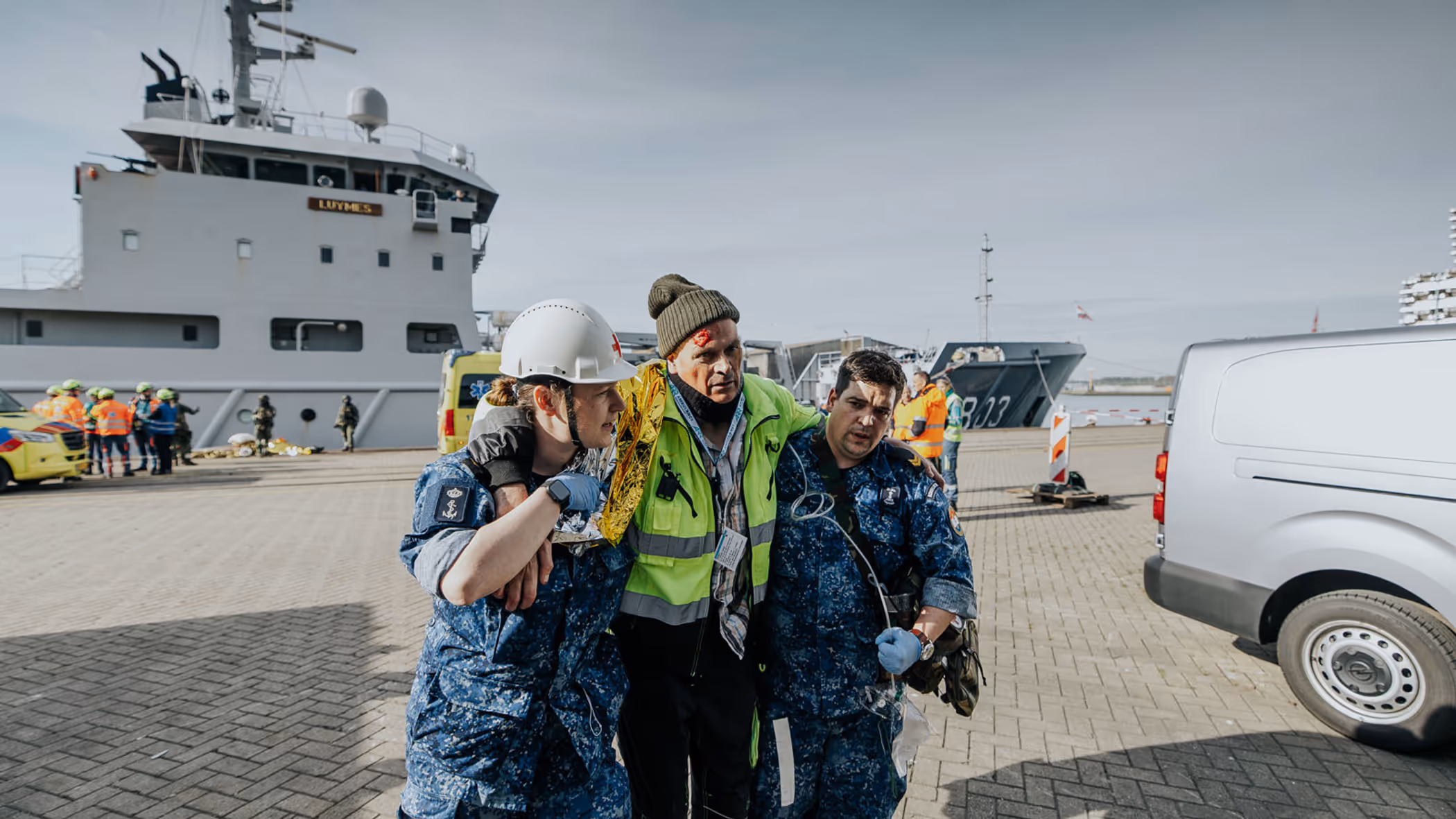 Een gewonde man wordt bij het lopen ondersteund door 2 militairen in boordtenue. Op de achtergrond is Zr.Ms. Luymes te zien.  