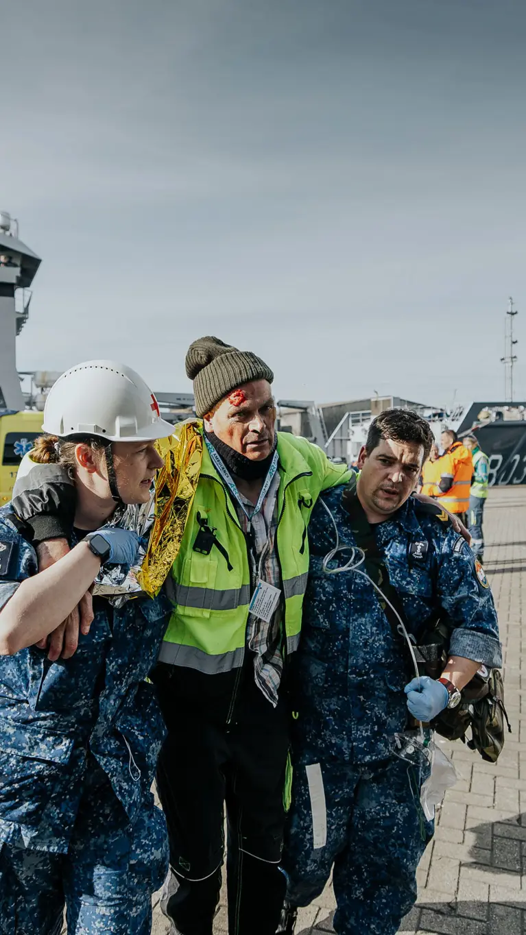 Een gewonde man wordt bij het lopen ondersteund door 2 militairen in boordtenue. Op de achtergrond is Zr.Ms. Luymes te zien.  