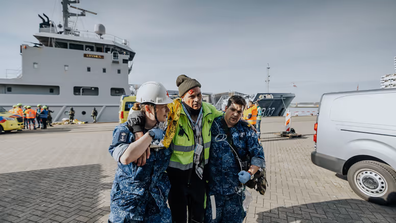 Een gewonde man wordt bij het lopen ondersteund door 2 militairen in boordtenue. Op de achtergrond is Zr.Ms. Luymes te zien.  