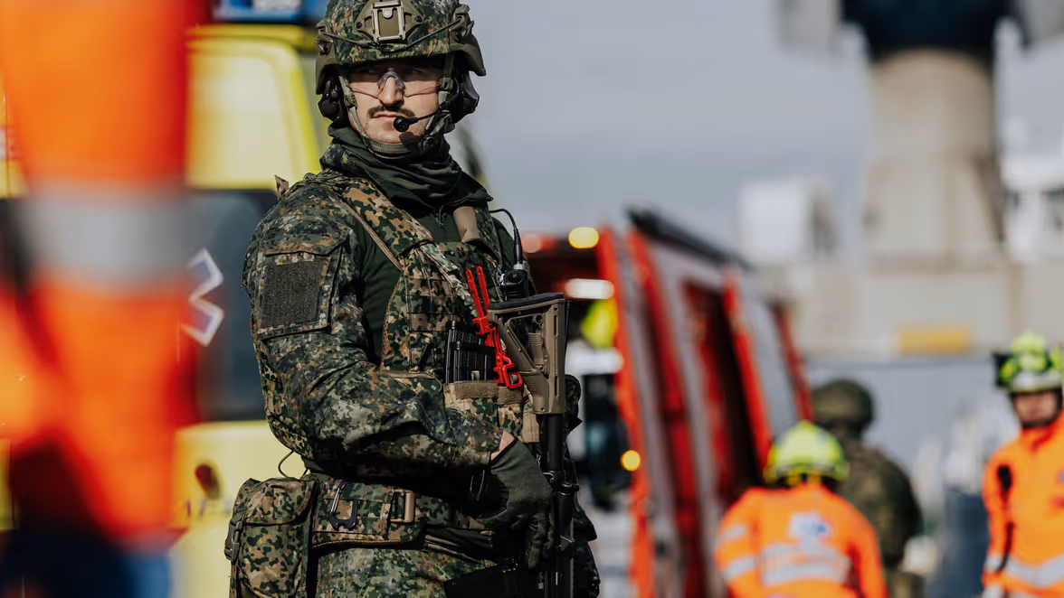 Een militair in groen vlekkenpak kijkt opzij. Achter hem staat personeel van de Brandweer. 