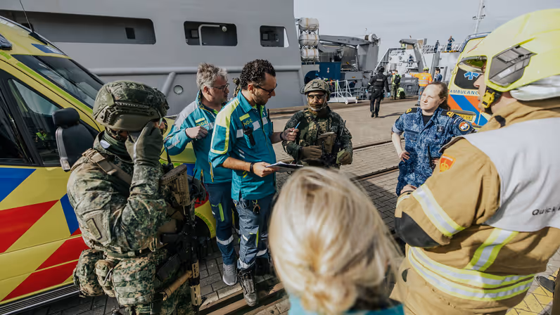 Een groep mensen staat in een cirkel te overleggen, onder hen militairen, ambulancepersoneel en Brandweerpersoneel. 