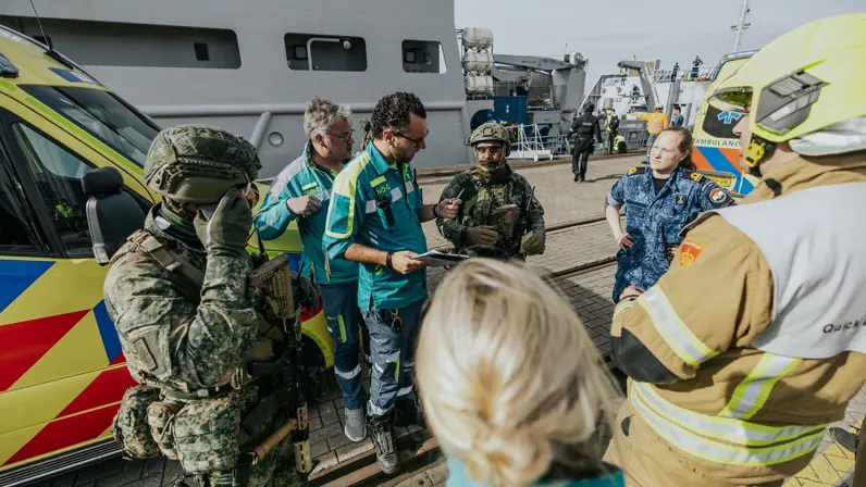 Een groep mensen staat in een cirkel te overleggen, onder hen militairen, ambulancepersoneel en Brandweerpersoneel. 