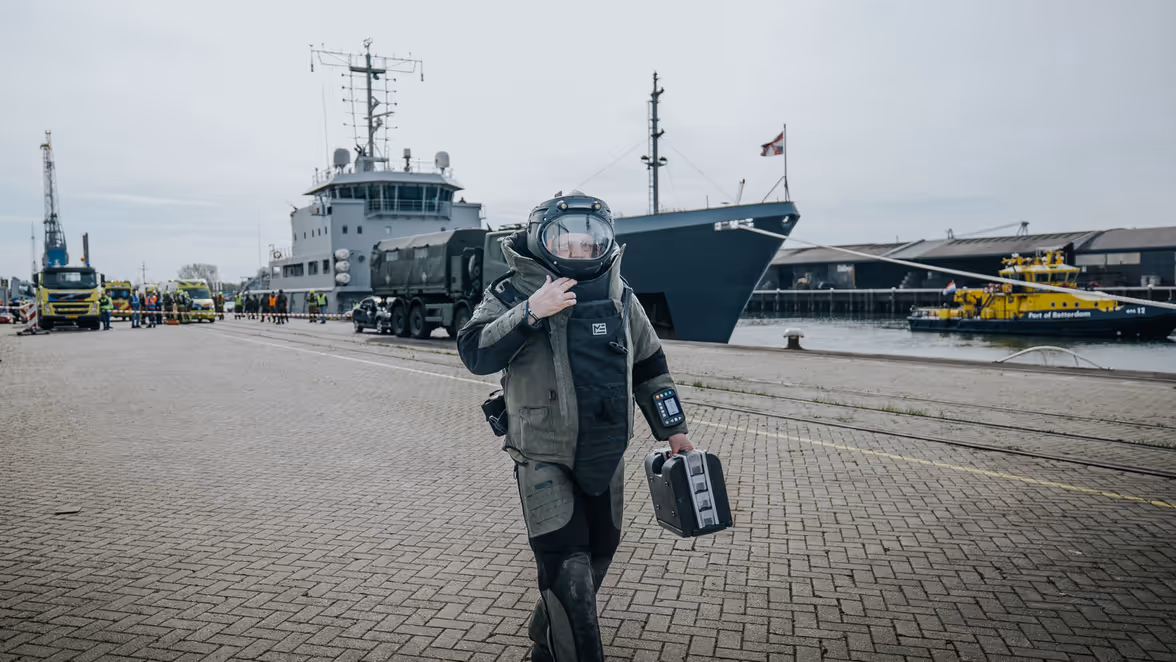 Een man in bompak loop over de kade. In zijn linkerhand draagt hij een kleine zwarte koffer. Achter hem is een schip te zien. 