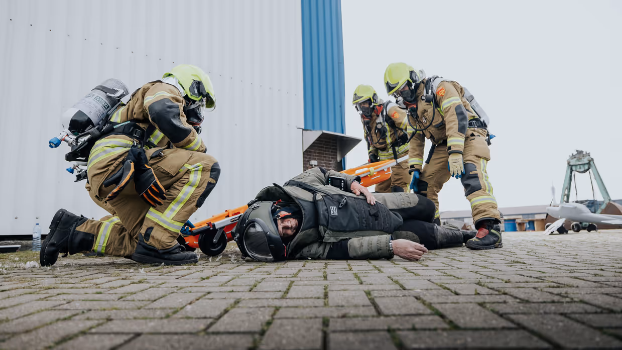 Alttekst = Een man in bompak ligt gewond op de grond. Hij wordt meegenomen door brandweerpersoneel.  