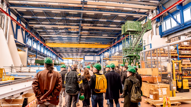 Groep mensen met groene helmen op loopt door hangar.