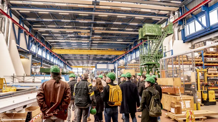 Groep mensen met groene helmen op loopt door hangar.