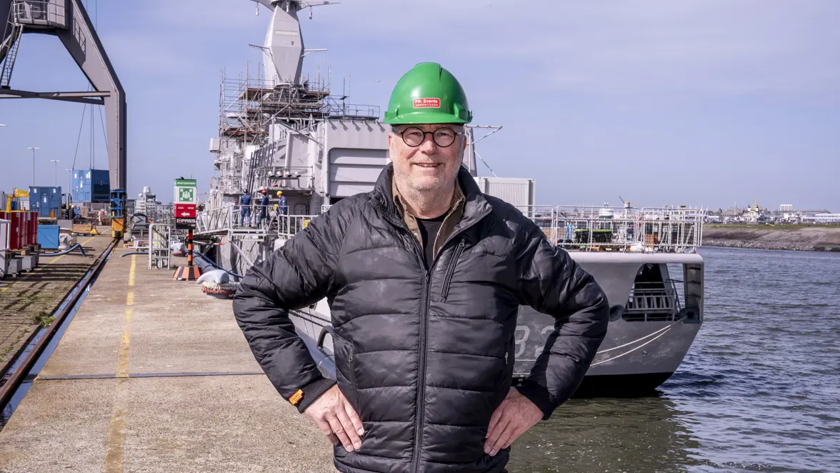 Man met groene helm op staat op kade voor marineschip.