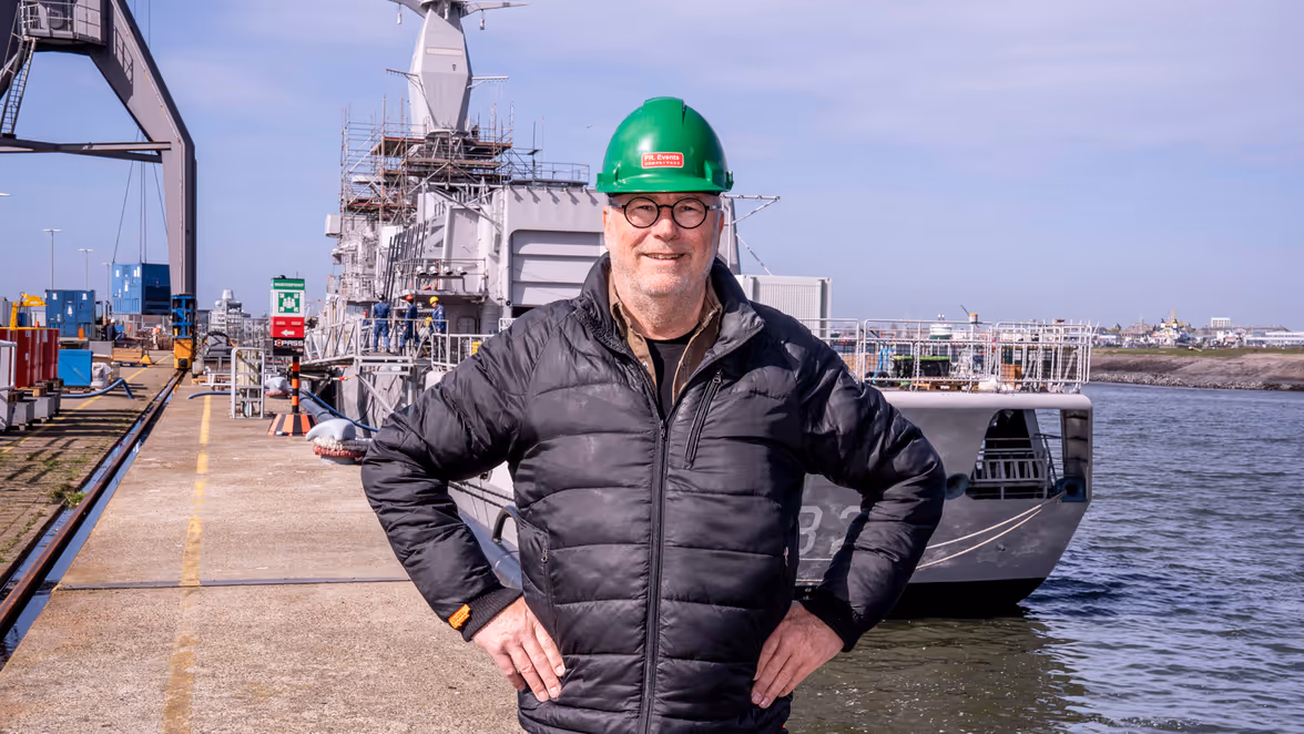 Man met groene helm op staat op kade voor marineschip.