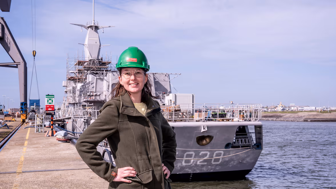 Vrouw met groene helm op staat op kade voor marineschip.