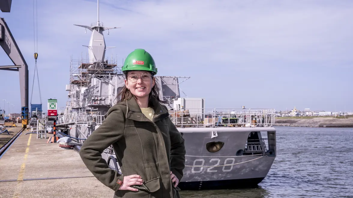 Vrouw met groene helm op staat op kade voor marineschip.