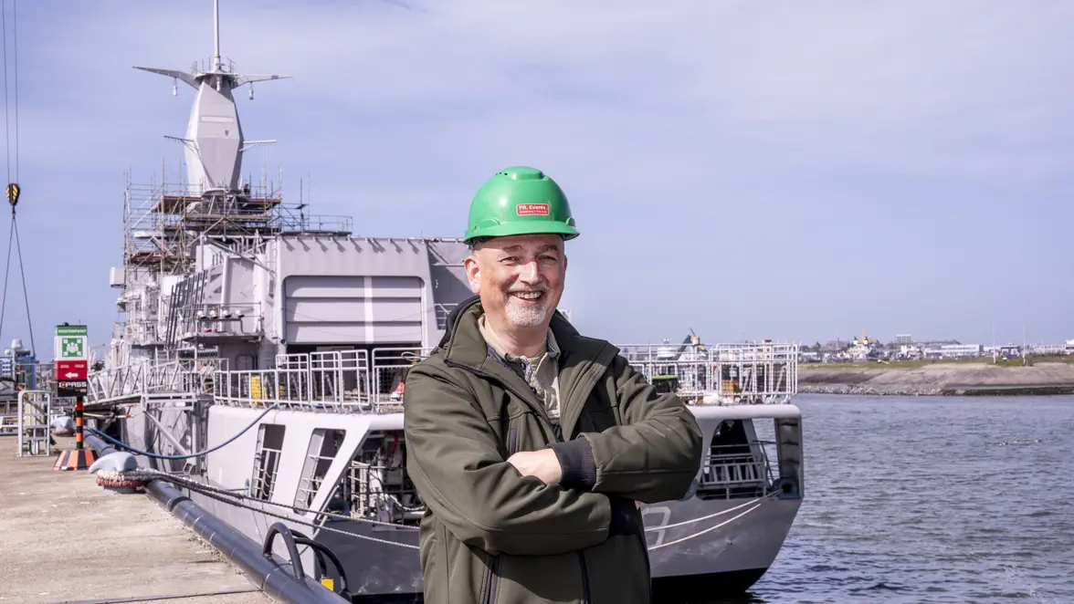 Man met groene helm op staat op de kade voor een marineschip.