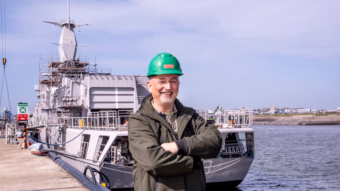 Man met groene helm op staat op de kade voor een marineschip.