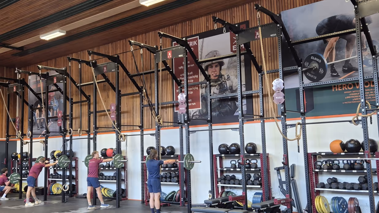 Een sportschool met krachttrainingsapparatuur. Er hangen gewichten, touwen en stangen. Vier mensen zijn aan het trainen, en de vloer is bedekt met rubberen matten.