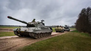 Een tank en gepantserde voertuigen rijden over een onverhard pad door een groen, open veld onder een bewolkte lucht. Rechts: Een zwaar militair voertuig met dubbele kanonnen en radar rijdt over een onverhard pad, gevolgd door andere legervoertuigen in een groen veld.