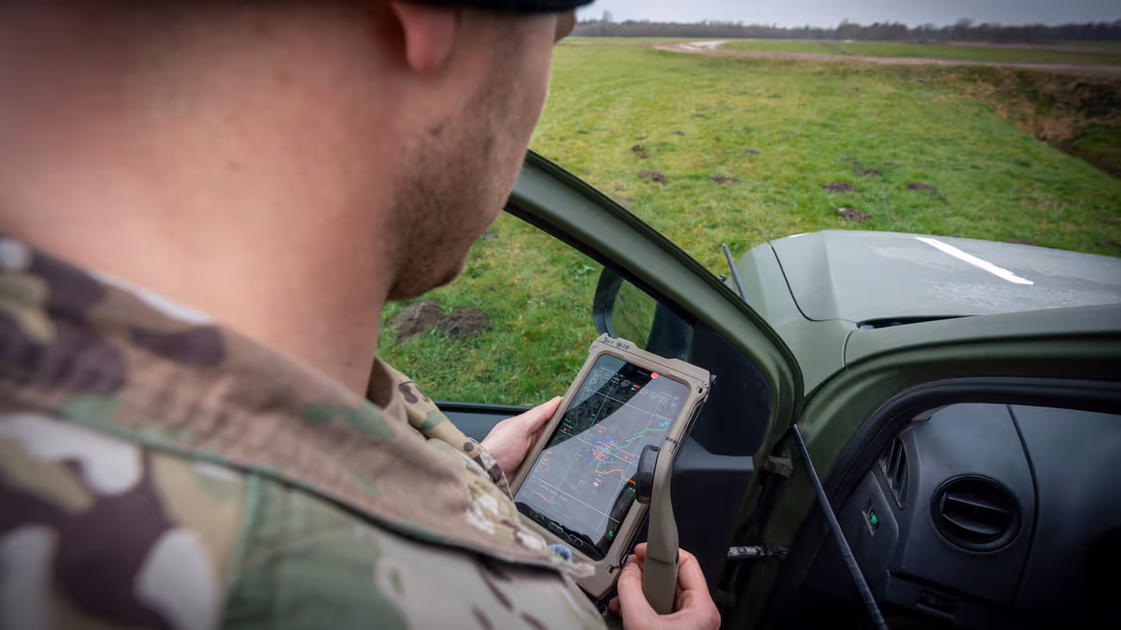 Een persoon in militair tenue houdt een tablet met kaart en routes vast in een voertuig, buiten is groen grasveld en een weg zichtbaar. Rechts: Een militair uniform met Nederlandse vlag en het woord 'ATTACK' op een rood label.