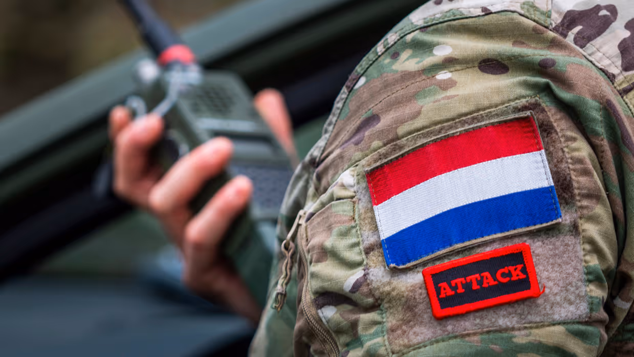 Een persoon in militair tenue houdt een tablet met kaart en routes in een voertuig, buiten is groen grasveld en een weg zichtbaar. Rechts: Een militair uniform met Nederlandse vlag en het woord "ATTACK" op een rood label  houdt een radio vast. 