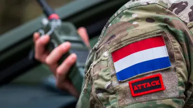 Een persoon in militair tenue houdt een tablet met kaart en routes in een voertuig, buiten is groen grasveld en een weg zichtbaar. Rechts: Een militair uniform met Nederlandse vlag en het woord "ATTACK" op een rood label  houdt een radio vast. 