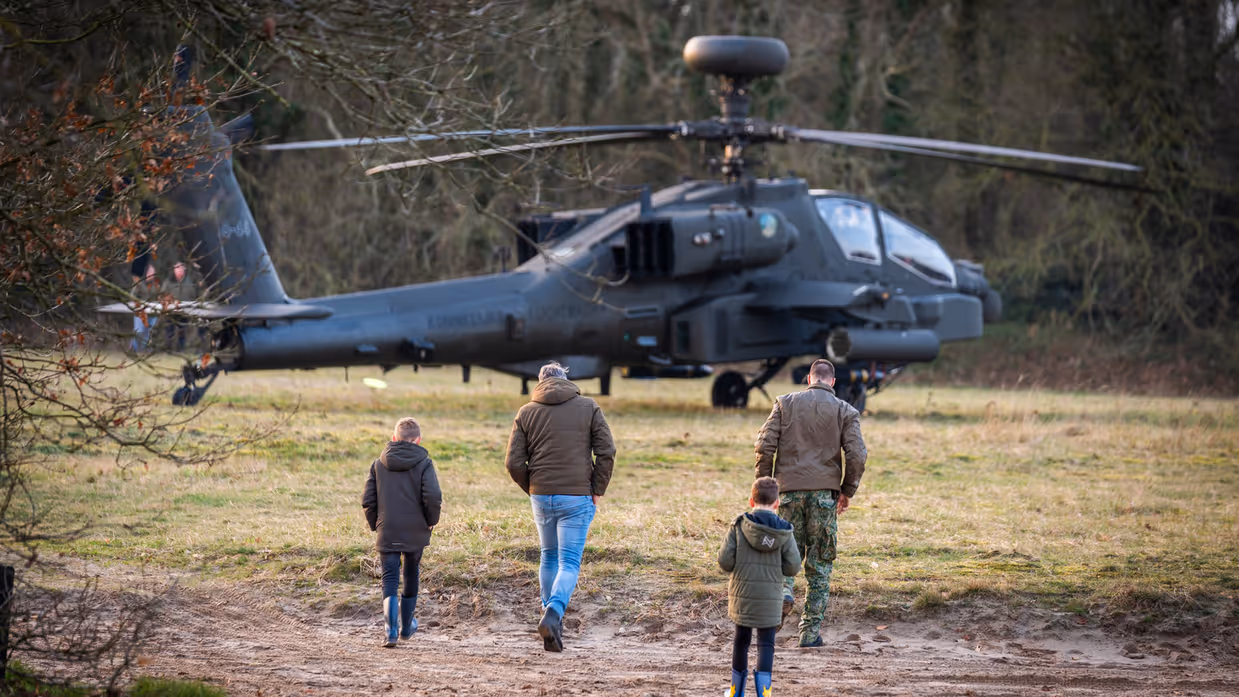 Vier personen, onder wie twee kinderen, lopen naar een militaire helikopter die op een grasveld tussen bomen staat geparkeerd. Rechts: Een militair in vliegtenue met helm en rugzak loopt weg van een grote, grijze militaire helikopter, omringd door bomen en gras. Op de achtergrond zijn kinderen te zien. 